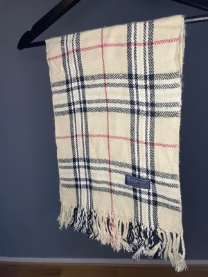 Burberry Halsduk Beige - Klassisk halsduk från Burberry i beige med rutigt mönster i svart, vitt och rött. Gjord i 100% lammull och har snygga fransar i ändarna. Perfekt accessoar för att lyfta din outfit med en tidlös brittisk vibe.