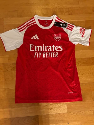 Arsenal Calafiori 33 matchtröja Adidas S - Riktigt snygg Arsenal matchtröja från Adidas i slim fit, med Calafiori och nummer 33 på ryggen. Röd kropp med vita korta ärmar och vita detaljer, samt diskret mönster i tyget. Officiell Premier League-tryck och Aeroready-material för bästa komfort.