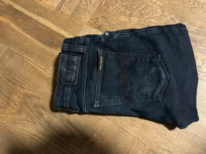 Svarta Nudie Jeans Skinny 29/32 - Säljer ett par svarta skinny jeans från Nudie Jeans i storlek 29/32. De har klassisk femficksdesign, svarta sömmar och Nudie-logga på bakfickan. Jeansen är tillverkade i stretchig denim för en tight passform och har knappgylf.