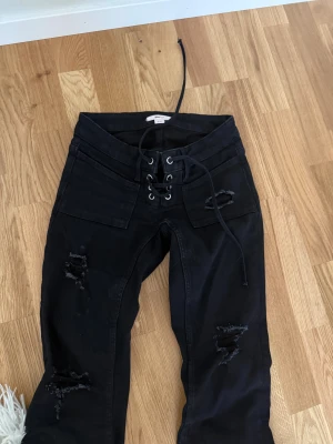 Svarta jeans med snörning och slitningar - Svarta jeans från Gina Tricot med cool snörning framtill och slitna detaljer på benen. Låg midja och raka bakfickor. Jeansen har en bootcut passform och är tillverkade i bomullsmix med stretch. Perfekta för en edgy streetstyle-look. TALL!!
