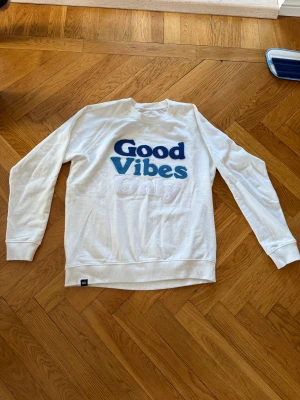 Vit sweatshirt från Dedicated, M - Vit sweatshirt från Dedicated med broderad text 'Good Vibes Only' i blått och vitt på bröstet. Tröjan är tillverkad av 100% ekologisk och Fairtrade-certifierad bomull. Klassisk rund hals och långa ärmar, perfekt för chill dagar. Ny pris runt 1 000 kr