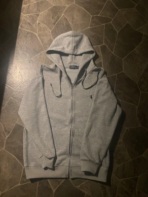 Grå hoodie från Polo Ralph Lauren - Snygg grå zip hoodie från Ralph lauren, säljer pågrund av storleken. Tröjan har inga defekter och knappt använd, Storlek L         Hör av er om ni är intresserade 