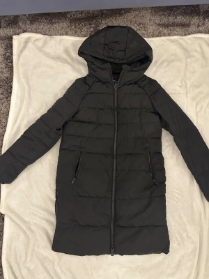 Svart lång pufferjacka Vero Moda - Svart lång pufferjacka från Vero Moda med huva och två sidofickor med dragkedja. Jackan har quiltad design och hel dragkedja framtill. Perfekt för kalla dagar och har en stilren look som funkar till det mesta.