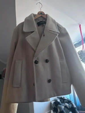 Beige kappa från Vero Moda - Snygg beige kappa från Vero Moda med bred krage och dubbelknäppning framtill. Jackan har två stora fickor och en rak, lite kortare passform som ger en clean och modern look. Perfekt för dig som gillar stilrena och klassiska plagg. Passar även Xs och S.