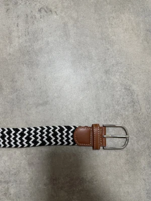 Grish bälte - Snyggt bälte med svartvitt zigzag-mönster och detaljer i brunt skinn. Spännet är i silverfärgad metall och bältet har en flätad design som ger en cool och modern look. Perfekt accessoar för att lyfta vilken outfit som helst.