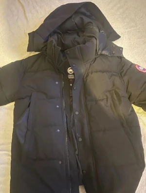 Svart dunjacka från Canada Goose - Svart dunjacka från Canada Goose med klassisk logga på ärmen. Jackan har huva, dragkedja och knappar framtill samt flera fickor. Perfekt för kalla dagar och har en clean, stilren look som funkar till allt. Fylld med dun och fjädrar för extra värme.