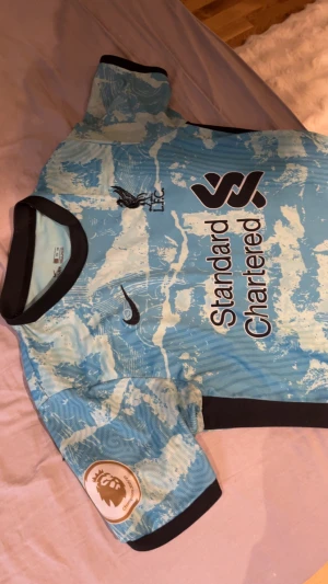 Liverpool FC blå bortatröja Nike - Liverpool FC bortatröja från Nike med coolt blått och vitt mönster, svart krage och detaljer. Tryck med Standard Chartered och Premier League-märke på ärmen. Dri-FIT material som andas, perfekt för match eller träning.