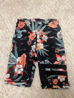 Blommiga träningsshorts från Stronger🌸 - Snygga träningsshorts från Stronger i modellen Jungle Fusion. Svart botten med färgglatt blommigt mönster i orange, vitt och grönt. Tight passform och hög midja, perfekta för gymmet eller löpning. Ord pris 499kr