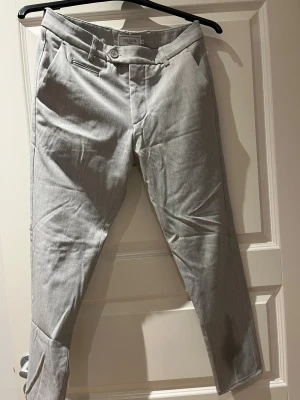 Slim fit kostymbyxor från Les Deux - Snygga ljusgrå kostymbyxor från Les Deux i slim fit-modell. Byxorna har klassiska pressveck, bälteshällor, framfickor och bakfickor med knapp. Perfekta för en clean och stilren look. Materialet är mjukt och känns lyxigt.