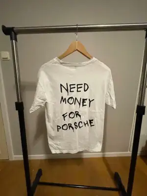 Vit t-shirt i bomull med svart tryck på ryggen där det står 'NEED MONEY FOR PORSCHE' i handskriven stil. Klassisk passform och rund halsringning, perfekt för dig som gillar statement-plagg.