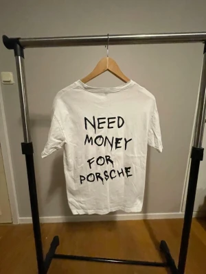 Need Money For Porsche T-shirt - Vit t-shirt i bomull med svart tryck på ryggen där det står 'NEED MONEY FOR PORSCHE' i handskriven stil. Klassisk passform och rund halsringning, perfekt för dig som gillar statement-plagg.
