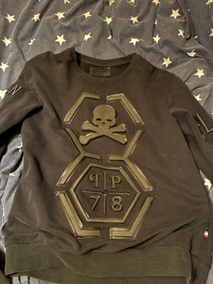 Svart Philipp Plein sweatshirt  - Svart sweatshirt från Philipp Plein med stort dödskalle- och benkorsmärke framtill, samt PP 78-logga i upphöjt material. Tröjan har ribbade muddar och lång ärm, tillverkad i 100% bomull. Snygga detaljer på ärmen och ryggen med PP-logga. Storlek L/M