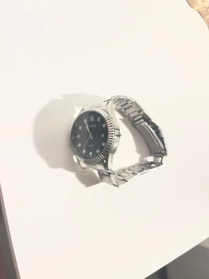 Silverfärgad Relock klocka med svart urtavla - Stilren klocka från Relock med silverfärgat metallarmband och svart urtavla. Urtavlan har markerade timmar med små stenar och datumvisning. Rund boett med räfflad kant ger en cool vibe. Perfekt accessoar för dig som gillar klassisk design med lite bling.