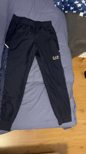 Svarta EA7 Emporio Armani byxor - Snygga svarta byxor från EA7 Emporio Armani med elastisk midja och muddar vid benslut. Byxorna har diskret logga på ena benet och mönstrade sidopaneler. Perfekta för en sportig och trendig look. Kan gå ner i pris vid snabbaffär