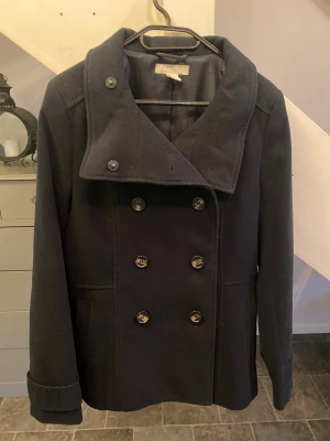 Svart rock från H&M, strl 40 -  Svart rock från H&M i storlek 40. Jackan har dubbelknäppning med sex knappar framtill, bred krage och knappdetalj bak. Tillverkad i mjukt ullmaterial och har långa ärmar. 