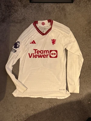 Manchester United 2023/24 tredje tröja player edition - Manchester United bortatröja med Mainoo 37, långärmad modell från Adidas. Vit med röda detaljer, klubbmärke och sponsortryck. Premier League-märke på ärmen och klassisk V-ringning. Tillverkad i lätt funktionsmaterial (HEAT.RDY).