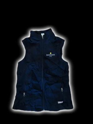 Svart fleeceväst Backroads Five från Patagonia  - Svart fleeceväst från Backroads Five med hög krage och broderad logga med gul stjärna på bröstet. Västen har två praktiska fickor med dragkedja framtill och hel dragkedja. Perfekt för lager-på-lager och chill stil.