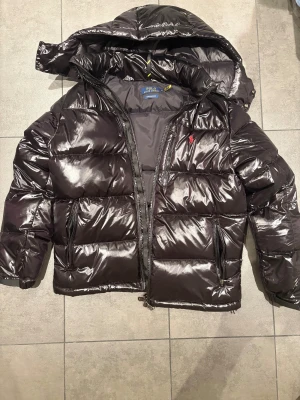 Svart pufferjacka Polo Ralph Lauren - Säljer en svart, glansig pufferjacka från Polo Ralph Lauren i storlek M. Jackan har huva, dragkedja framtill och två sidofickor med dragkedja. Klassisk quiltad design och röd Polo-logga på bröstet. Perfekt för kalla dagar och riktigt snygg streetstil. 