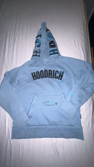Ljusblå Hoodie från Hoodrich M - Snygg ljusblå hoodie från Hoodrich i storlek M med stor svart logga på bröstet och coola tryck på huvan. Tröjan har känguruficka och text på ryggen. Mjuk insida i fleece och ribbade muddar. Perfekt för dig som gillar streetwear och vill sticka ut.