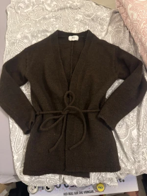 Mörkbrun kofta med bälte från Gina Tricot - Mysig mörkbrun kofta från Gina Tricot i ullblandning. Modellen är omlott med knytband i midjan och har långärmade ärmar. Perfekt för lager-på-lager och chill dagar. Enkel, stilren och passar dig som gillar klassiska vibes.