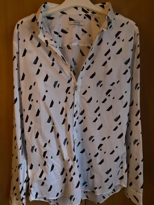 Vit skjorta med svart mönster Premium - Snygg vit skjorta från Premium med svart och grått abstrakt mönster. Skjortan har klassisk krage, knappar framtill och långa ärmar. Perfekt för dig som gillar stilrena plagg med en twist. Materialet känns lätt och bekvämt mot huden.