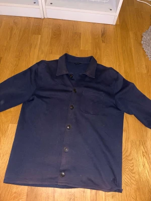 Mörkblå overshirt från Riley - Snygg mörkblå overshirt från Riley i storlek M. Skjortan har klassisk krage, knäppning med svarta knappar hela vägen och en bröstficka. Materialet är mjukt och känns som bomull, perfekt för lager-på-lager. Långärmad och stilren design.