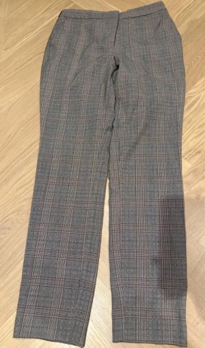 Grå rutiga kostymbyxor - Snygga grå kostymbyxor med klassiskt rutig mönster och raka ben. Byxorna har en stilren design med diskreta fickor och passar perfekt till en dressad look. Materialet känns mjukt och har en lätt struktur som ger extra stilpoäng.
