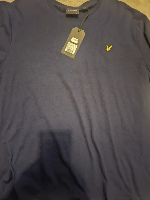 Blå t-shirt från Lyle & Scott - Snygg marinblå t-shirt från Lyle & Scott med klassisk rund hals och korta ärmar. På bröstet sitter den ikoniska gula fågeln som broderad detalj. T-shirten är gjord i mjuk bomull och har en stilren, enkel design som funkar till allt.