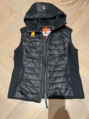 Svart quiltad dunväst Parajumpers S - Svart dunväst från Parajumpers i storlek S med quiltad framsida, huva och coolt PJS-märke på huvan. Västen har dragkedja framtill och detaljer i mesh på sidorna. Perfekt för lager-på-lager och streetstyle.