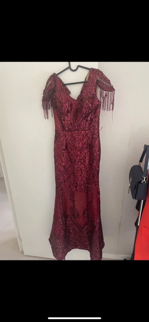 Vinröd glittrig långklänning med fransar - Vinröd långklänning med glittriga paljetter och v-ringning. Klänningen har offshoulder-ärmar med fransdetaljer och en figurnära passform som ger en snygg siluett. Perfekt för bal eller fest där du vill sticka ut. Materialet är glittrigt och skimrande för extra wow-effekt. De även ett mjukt material under klänningen. Klänningen har använts 1 gång och tyvärr låsande lite pärlor, men klänning är i jätte bra skick.