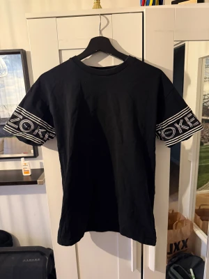 Svart Kenzo t-shirt med tryck - Svart t-shirt från Kenzo med coolt vitt tryck på ärmarna där det står 'KENZO PARIS'. Klassisk rund hals och rak passform. Materialet är mjuk bomull och ärmarna är korta med ett grafiskt mönster som sticker ut.