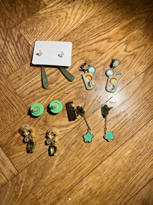 Mix av fem par örhängen i grönt - Säljer ett set med fem par örhängen i olika stilar och gröna nyanser. Inkluderar droppformade örhängen, gröna studs, cocktailglas med pärldetaljer, blommiga hängörhängen, glittriga örhängen med stenar och ett par med geometrisk design. Perfekt för dig som gillar att variera din look.