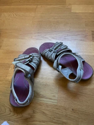 Beige sandaler från Ecco med lila sula - Säljer ett par beige sandaler från Ecco med justerbara remmar och lila innersula med Shockpad-dämpning. Sandalerna har en platt sula med mönstrad undersida för extra grepp. Perfekt för sommaren och långa promenader.