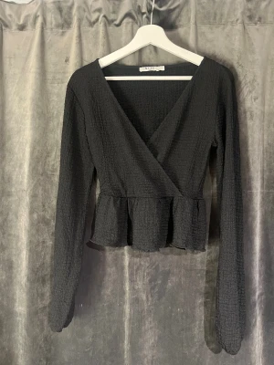 Svart omlottopp med volang från NA-KD - Säljer en svart omlottopp från NA-KD i storlek S. Toppen har långärmade ärmar, v-ringning och en snygg volangkant nedtill. Materialet är strukturerat och ger en cool textur. Perfekt att styla med jeans eller kjol för en trendig look.