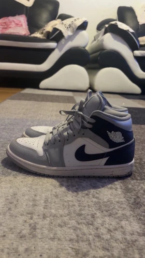 Nike Air Jordan 1 Mid grå/vit - Nike Air Jordan 1 Mid sneakers i grått och vitt med klassisk svart swoosh på sidan. Skorna har högt skaft, snörning och perforerad tå för extra ventilation. Tillverkade i skinn och syntetmaterial, med rund tå och platt sula. Perfekt för dig som gillar streetwear och ikoniska sneakers.