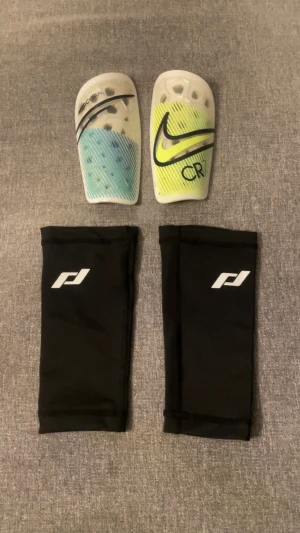 CR7 benskydd, sleeves - Säljer ett par Nike CR7 benskydd med tillhörande svarta sleeves. Benskydden har coolt randigt mönster i blått och gult med CR7-logga och Nike swoosh. Sleevesen är svarta med CR7- och Nike-logga i gult, perfekta för fotbollsträning eller match. Alla tillbehör säljs tillsammans, kan alltid vara bra att ha!