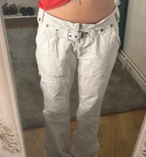 Vita cargopants med dragkedjor - Säljer ett par vita cargopants med coola detaljer som bälte i midjan, fickor med knappar och dragkedjor på benen. Byxorna har låg midja och en loose passform med raka ben. Perfekt för dig som gillar streetstyle och vill sticka ut.