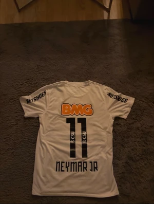 Santos Neymar Jr matchtröja Nike M - Säljer en äkta Santos FC fotbollströja från Nike, Neymar Jr #11. Tröjan är vit med svarta tryck, orange BMG-logga och klubbmärke på bröstet. Kortärmad, Dri-Fit-material för skön känsla. Netshoes-tryck på ärmarna och autenticitetsmärke. Perfekt för dig som älskar brasiliansk fotboll.
