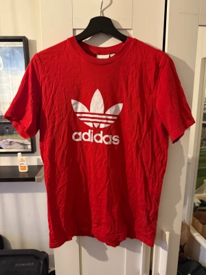 Röd Adidas t-shirt med logga - Säljer en röd t-shirt från Adidas med den klassiska vita trefoil-loggan tryckt på bröstet. T-shirten har rund hals och korta ärmar, tillverkad i mjuk bomull. Perfekt för dig som gillar sportig och enkel streetstyle.