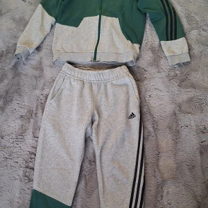 Adidassett stl 152 - Adidas hoodiejacka i grön och ljusgrå färg med svarta ränder på ärmarna. Jackan har huva, hel dragkedja och stora fickor framtill. Byxor med snörning i midjan och två fickor. Litet mörke från cykelkedja på ena benet, se bild. Materialet är mjuk polyester och bomull, perfekt för chill eller träning. Snygg tvåfärgad design med klassiska Adidas-loggan på bröstet.
