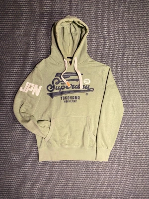 Superdry Hoodie - Snygg ljusgrön hoodie från Superdry med stort blått tryck på bröstet och vit JPN-text på ärmen. Klassisk känguruficka, vita snören i huvan och mjuk insida. Perfekt för dig som gillar streetstyle och japanska influenser. Om du undrar något är det bara att skriva🙌✅