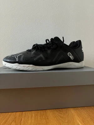 Svarta Puma fotbollsskor inomhus - Säljer ett par svarta inomhus fotbollsskor från Puma med vita detaljer och logga på både häl och tå. Skorna har snörning, ventilerande meshpaneler och en vit platt sula med greppigt mönster. Perfekta för futsal eller inomhusträning.