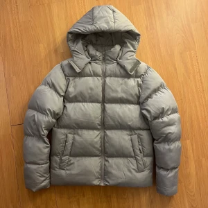 Grå pufferjacka från ASOS DESIGN - Säljer en grå pufferjacka från ASOS DESIGN med huva och dragkedja framtill. Jackan har en quiltad look, två sopiga fickor och är riktigt fluffig och varm. Perfekt för kalla dagar när du vill vara både chill och snygg. s/m