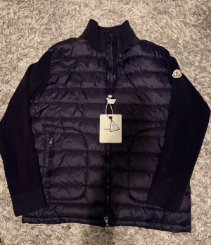 Mörkblå Moncler cardigan - Snygg mörkblå pufferjacka från Moncler med stickade ärmar och quiltad framsida. Jackan har hög krage, tvåvägsdragkedja och Moncler-logga på ärmen. Insidan har ett coolt serietryck och etikett. Har endas använt den 5 tillfällen. 