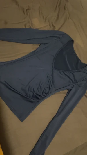 Mörkblå omlottopp med v-ringning - Säljer en mörkblå omlottopp med lång ärm och djup v-ringning. Toppen har en figurnära passform och är tillverkad i ett stretchigt, slätt material som ger en snygg siluett. Perfekt att styla med jeans eller kjol för en trendig look.