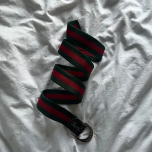 Randigt bälte från Gucci - Säljer ett snyggt bälte från Gucci med klassiska gröna och röda ränder. Bältet har en metallspänne med Gucci-loggan ingraverad. Perfekt accessoar för att lyfta din outfit med lite lyxig känsla.