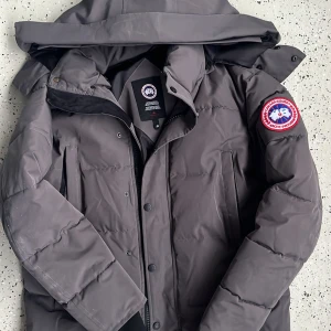 Mörkgrå Canada Goose dunjacka - Säljer en mörkgrå dunjacka från Canada Goose med avtagbar huva och pälsdetalj. Jackan har klassisk patch på ärmen, dragkedja och knappar framtill samt två sidofickor. Perfekt för kalla dagar med sin tjocka och quiltade design.