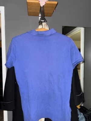 Blå pikétröja från Ralph Lauren - Snygg blå pikétröja från Ralph Lauren med klassisk krage och korta ärmar. Tröjan har två knappar vid halsen och en stor svart Polo-logga på bröstet samt en svart siffra på ärmen. Perfekt för dig som gillar sportig och stilren look.som ny