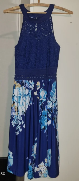 Blå blommig spetsklänning - Superfin blå klänning med spets upptill och blommigt mönster nertill. Klänningen har ärmlös design, rund halsringning och markerad midja med spetsdetaljer. Kjolen är utsvängd och har stora blommor i blått, vitt och gult. Perfekt för sommarens alla tillfällen!