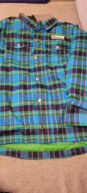  Snowboarjacka  - Snygg rutig overshirt från Burton i blått, grönt och svart med grå huva. Jackan har knappar framtill, två bröstfickor och dragkedja på insidan. Materialet är polyester och bomull, perfekt för dig som gillar streetstyle och vill sticka ut. Dragkedja är ihopsydd så jackan dras över huvudet. Därav priset. Passar ungdom 14=16 år